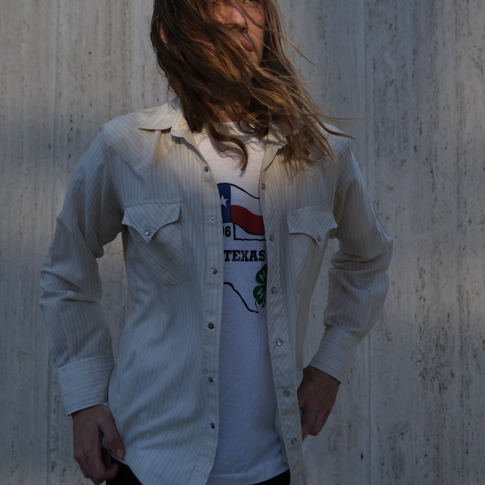 A musician with long hair wearing a striped shirt over a Texas-themed t-shirt, standing against a textured background. Barrett Avner
